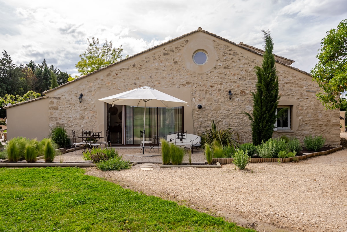 The exterior of the stone house features a large, round window and an inviting patio area with a white umbrella. A gravel pathway leads to the entrance, surrounded by lush greenery and decorative plants. Outdoor seating arrangements are visible adjacent to the house.