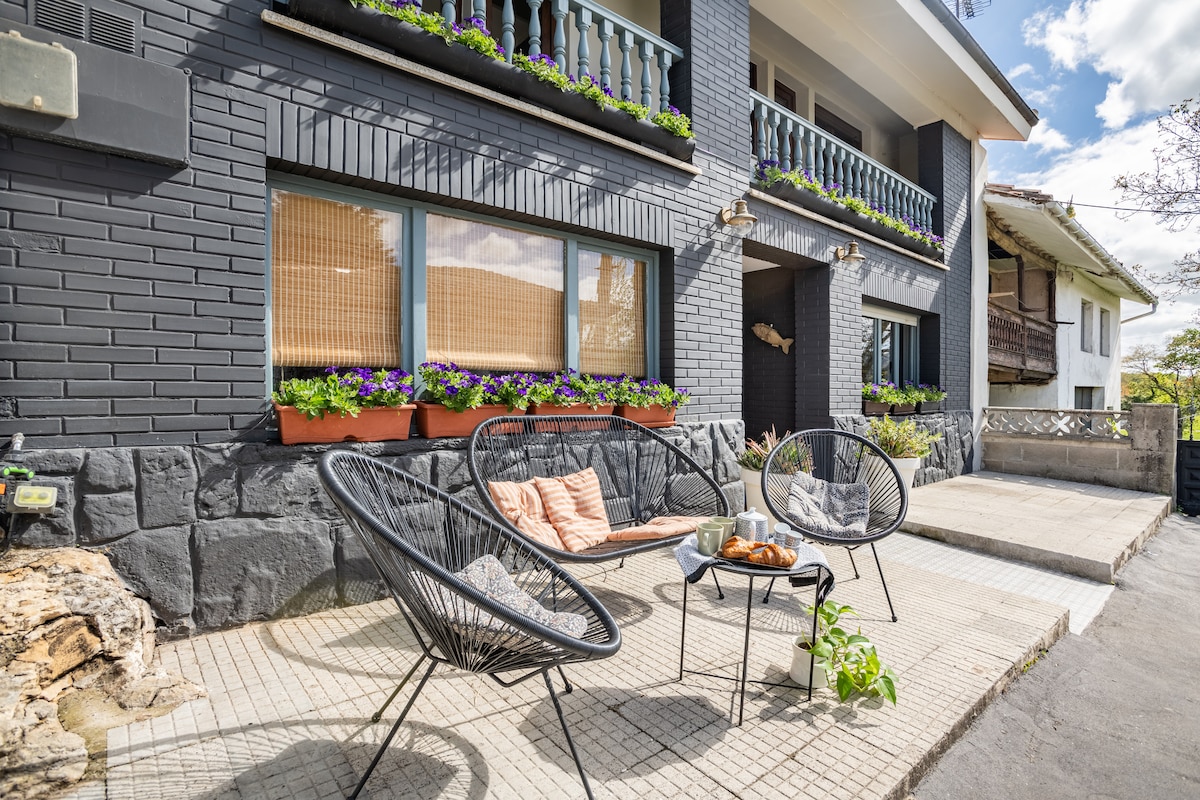 A charming outdoor area features black wicker chairs arranged around a small table, set with refreshments. Flower boxes adorned with vibrant blooms are positioned under the windows of the building, while the pathway is lined with textured tiles, contributing to a welcoming ambiance.