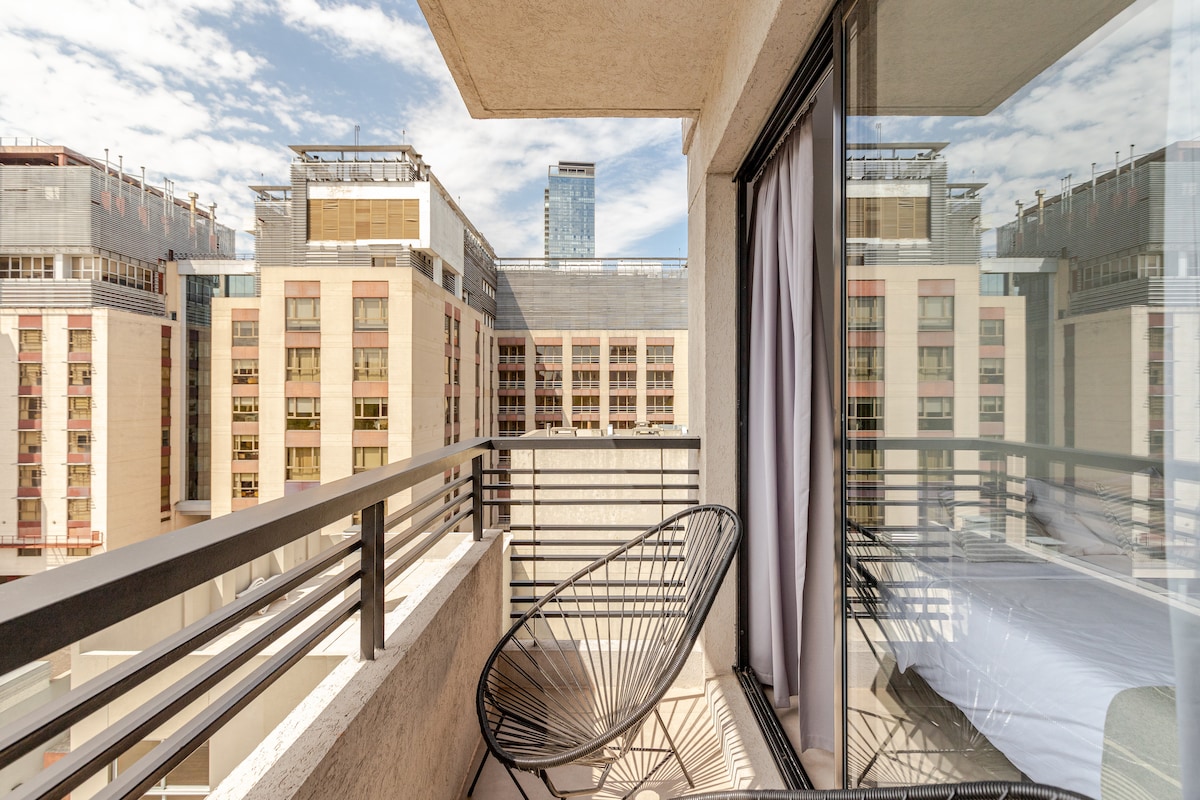 A private balcony offers an open view of the cityscape, framed by glass doors. A black chair sits on the balcony, providing a space to relax and enjoy the surroundings. The sky is partly cloudy, creating a bright atmosphere.