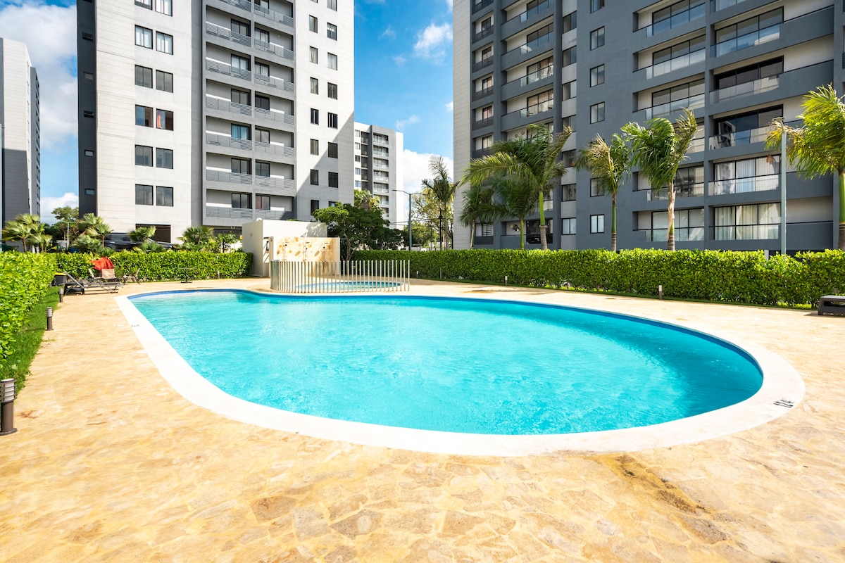 The outdoor area features a large swimming pool surrounded by lush greenery and palm trees. A stone pathway borders the pool, providing access to sun loungers. The apartment buildings create a backdrop against the clear blue sky.