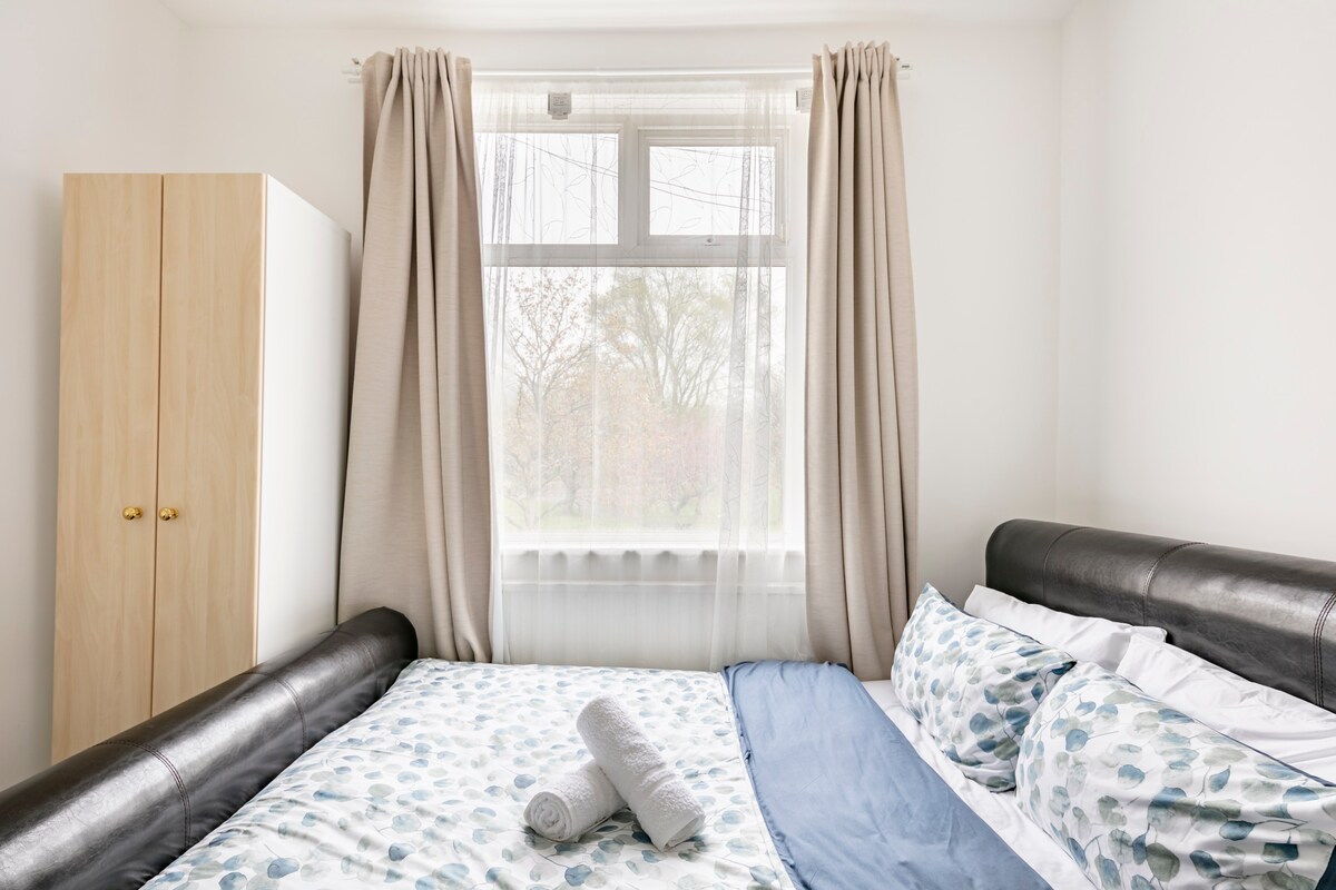 A double room features a spacious bed with patterned bedding and rolled towels atop it. Natural light enters through a large window with light curtains. A wardrobe stands next to the bed, enhancing the room's functionality.