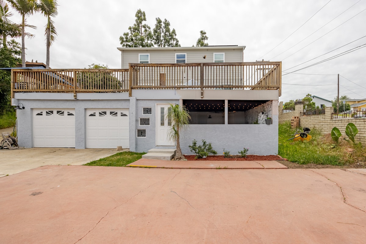 The exterior features a two-story home with a wooden deck above and a private entrance to the guest suite below. The garage doors offer ample parking space, and the garden area includes shrubs and decorative stones, creating a welcoming outdoor space.