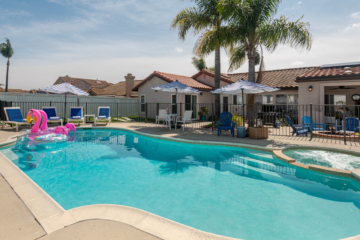 A clear blue swimming pool is surrounded by tropical palm trees and outdoor lounge chairs in a spacious backyard. Colorful umbrellas provide shade over the seating areas, while a jacuzzi sits adjacent to the pool. The inviting space is designed for relaxation and enjoyment.