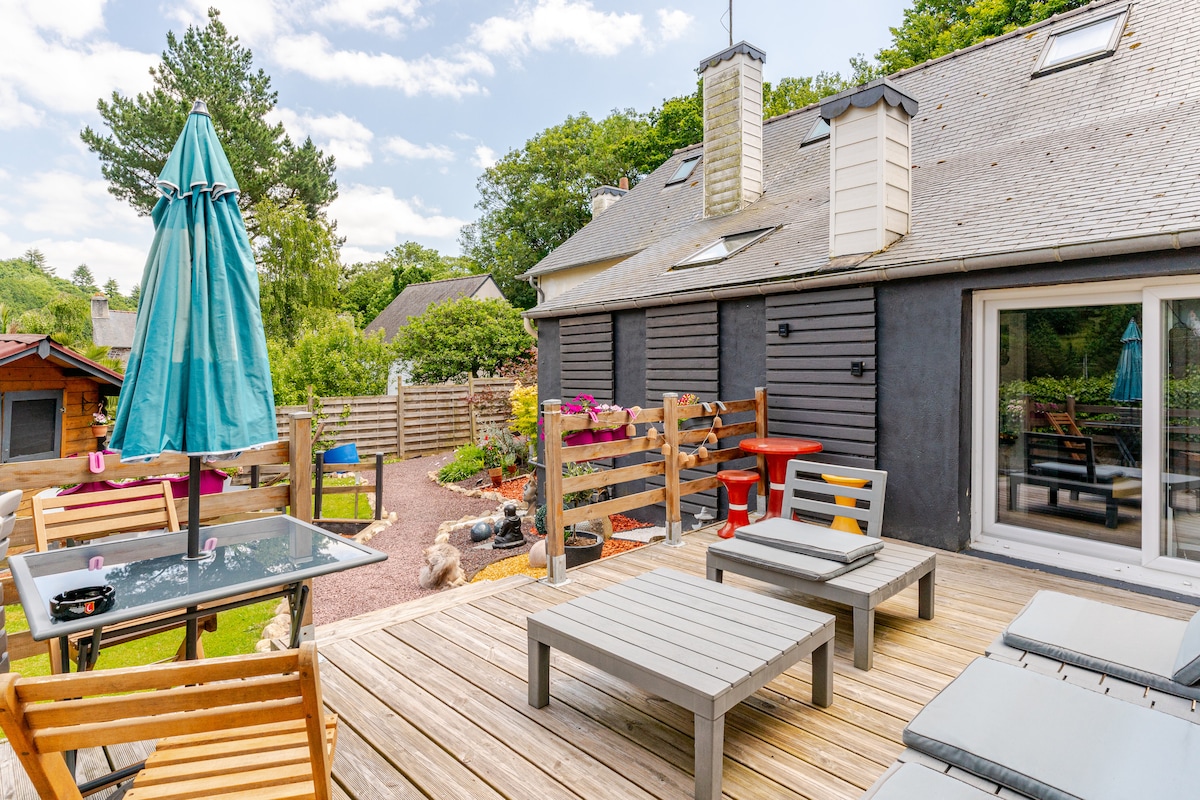 A spacious outdoor terrace is presented with wooden furniture, including a table and lounge chairs. A turquoise umbrella provides shade, while planters add greenery. The surrounding area features a wooden fence and lush trees, creating a peaceful atmosphere for relaxation.