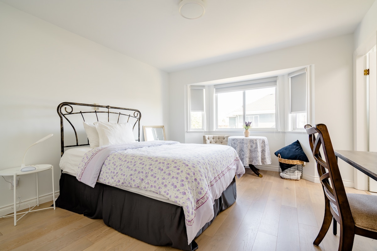 A bright bedroom features a decorative iron bed with linens adorned in floral patterns. Natural light enters through a large window seat, highlighting the light-colored wooden floor. A table and chair are arranged near the window, offering a space for work or relaxation.