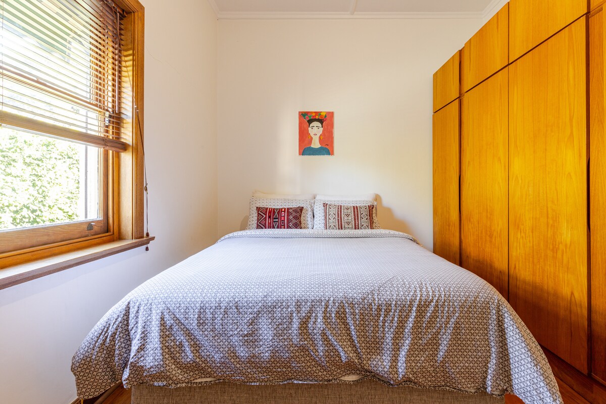 A cozy bedroom is furnished with a spacious bed adorned with patterned linens. Two decorative pillows rest against the headboard. A vibrant piece of artwork is mounted above the bed. Natural light filters through the window, illuminating the wooden cabinetry along the wall.