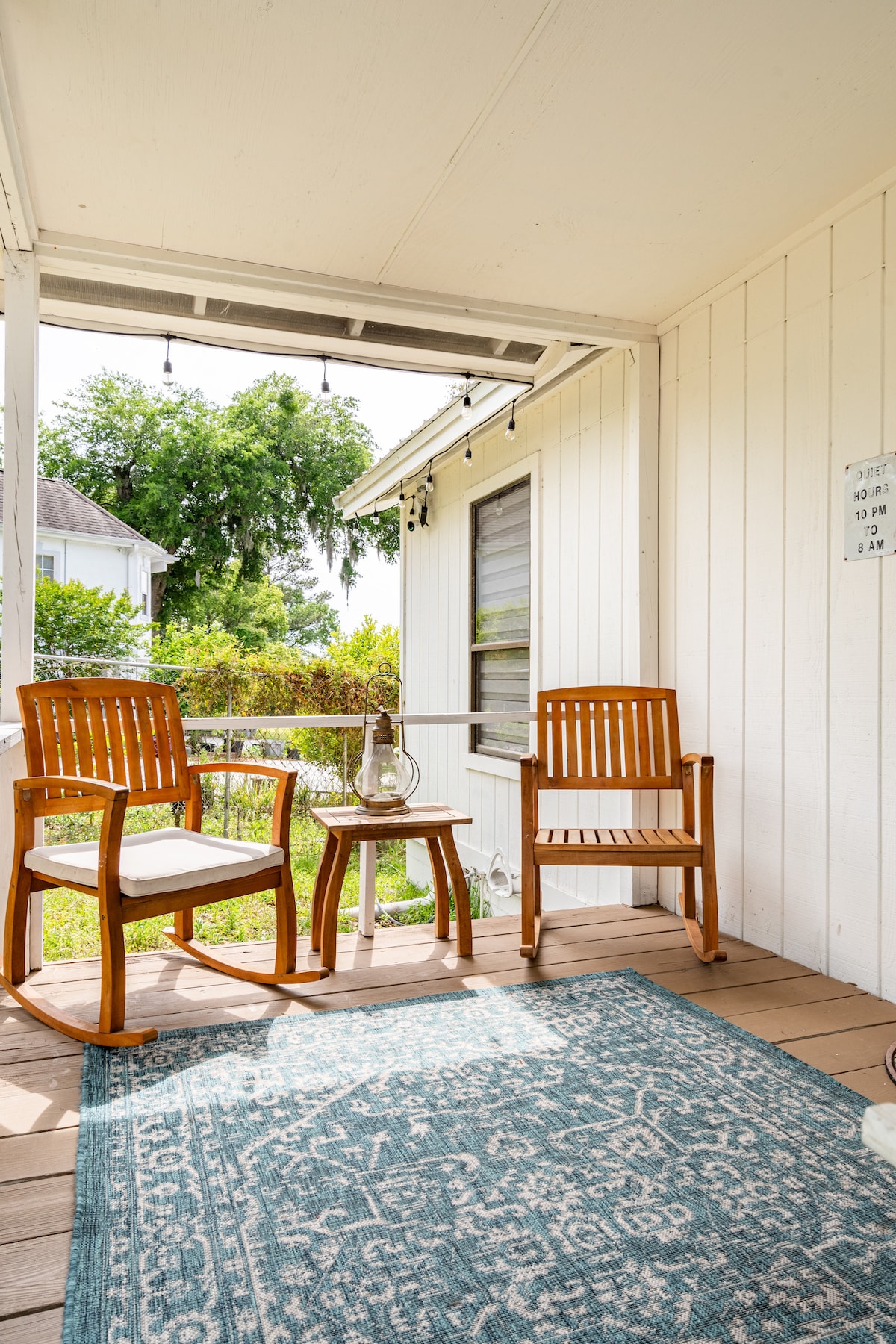 A welcoming porch area features two wooden chairs and a small round table, inviting relaxation. A decorative rug adds a touch of warmth beneath. Greenery surrounds the space, while string lights enhance the ambiance, providing a cozy spot to enjoy the outdoors.