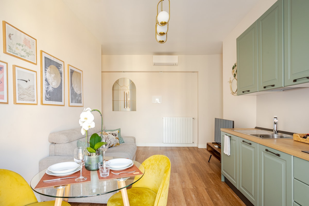 A cozy dining area features a wooden table set for two, complemented by yellow chairs. A stylish kitchen space is visible in the background, with green cabinets and countertop appliances. Light is provided by a contemporary pendant light, enhancing the warm atmosphere.