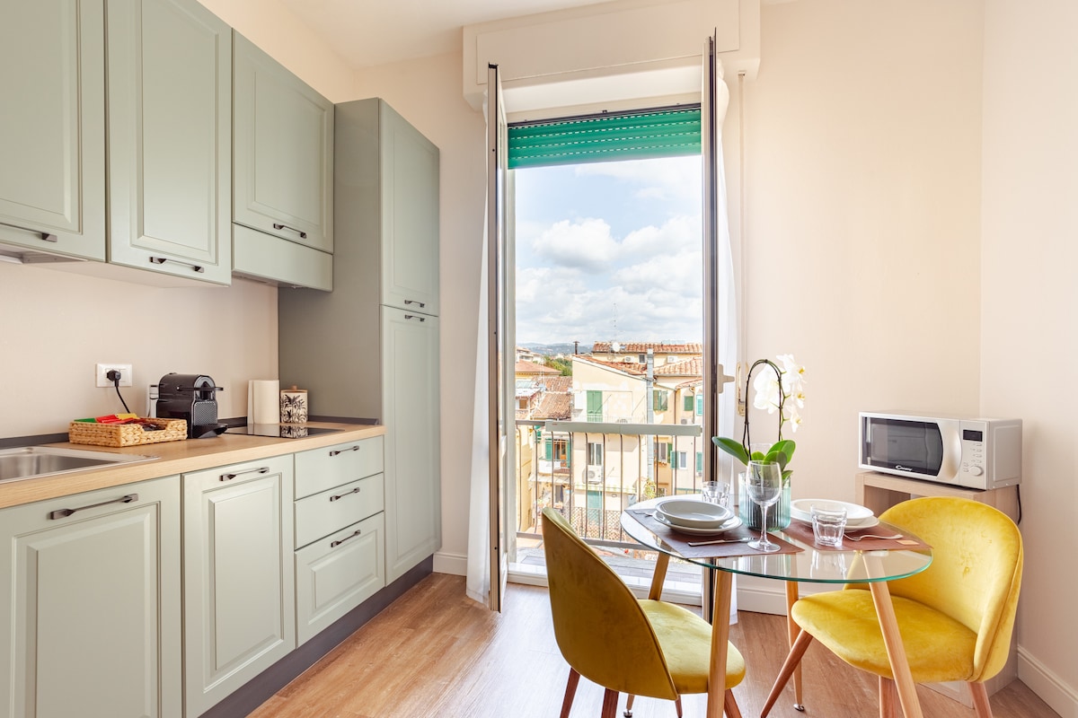 A well-appointed kitchenette is featured, displaying light green cabinetry and a countertop. A dining table with two yellow chairs is positioned by a window, which offers views of the surrounding rooftops. Natural light fills the space, enhancing its bright and inviting feel.