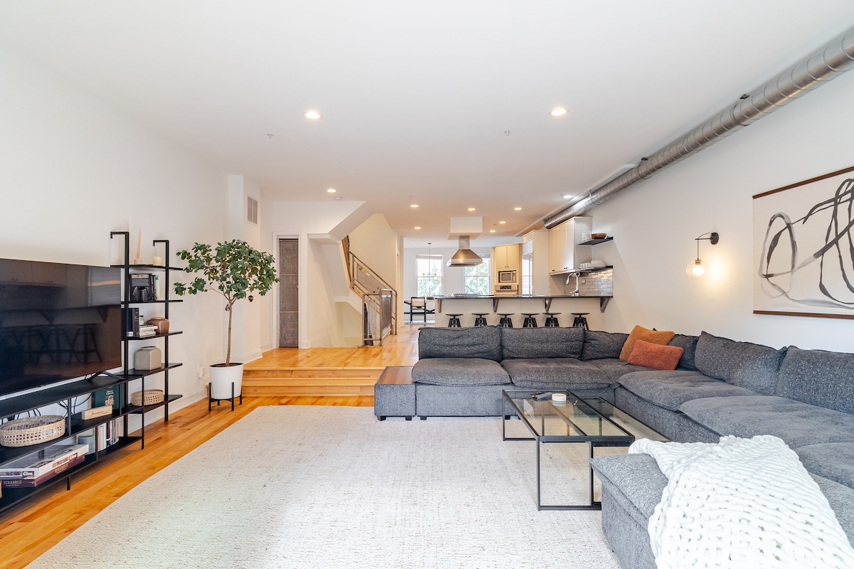 An open concept living area is highlighted, featuring a modular pit sofa set on a light rug. A sleek coffee table is positioned in front of the sofa. In the background, the kitchen and dining area are visible, with modern fixtures and a staircase leading to the upper level.