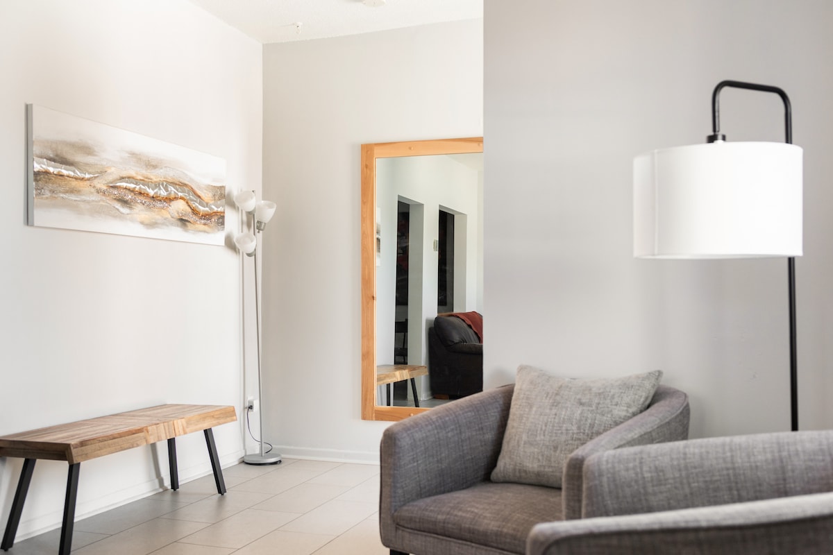 A light-filled living area features a modern gray armchair and a wooden bench against a neutral wall. A standing lamp adds a soft glow, while a large mirror reflects the space. An abstract wall art piece adds visual interest.