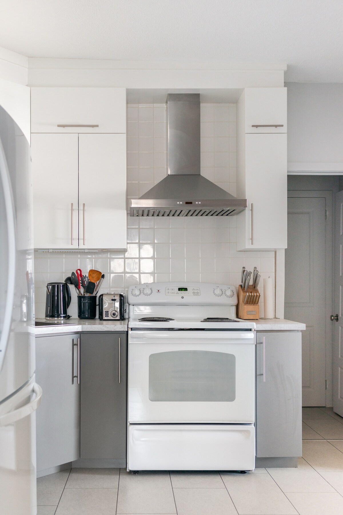 The kitchen features modern cabinetry in light and dark tones, with a sleek white stove and silver hood. Kitchen appliances are neatly arranged, including a coffee maker and toaster. A tiled backsplash complements the countertop, enhancing the functional cooking space.