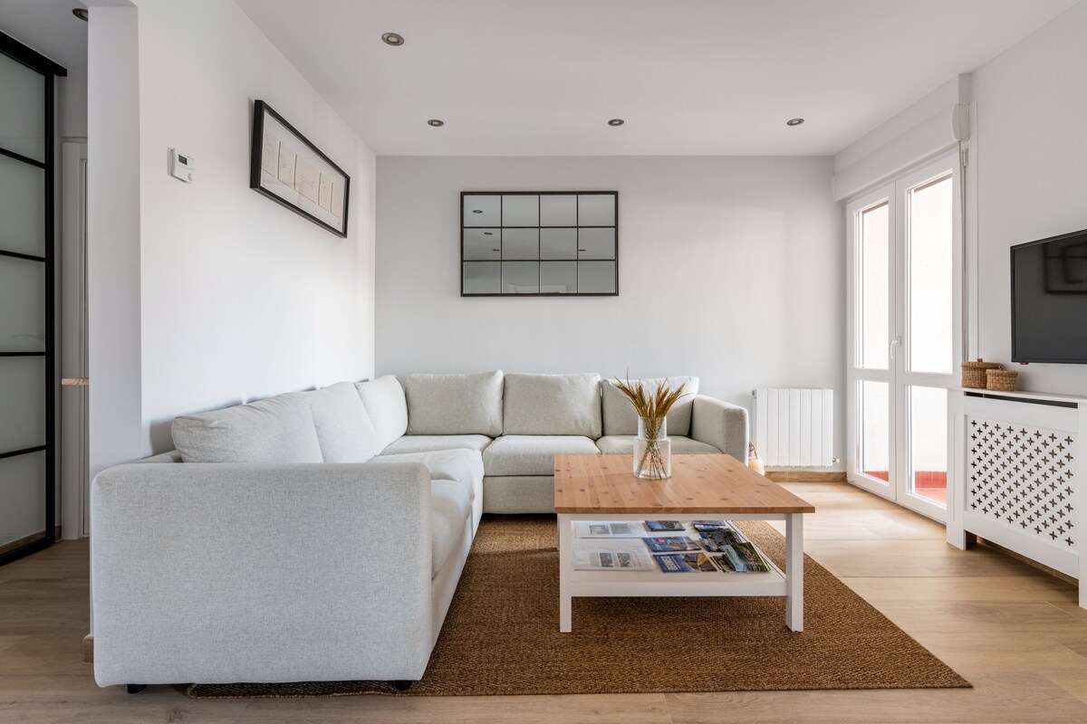 A bright living room features a light-colored sectional sofa situated around a wooden coffee table. A decorative plant is placed on the table, and large windows allow natural light to illuminate the space. Simple wall art is displayed, enhancing the modern decor.