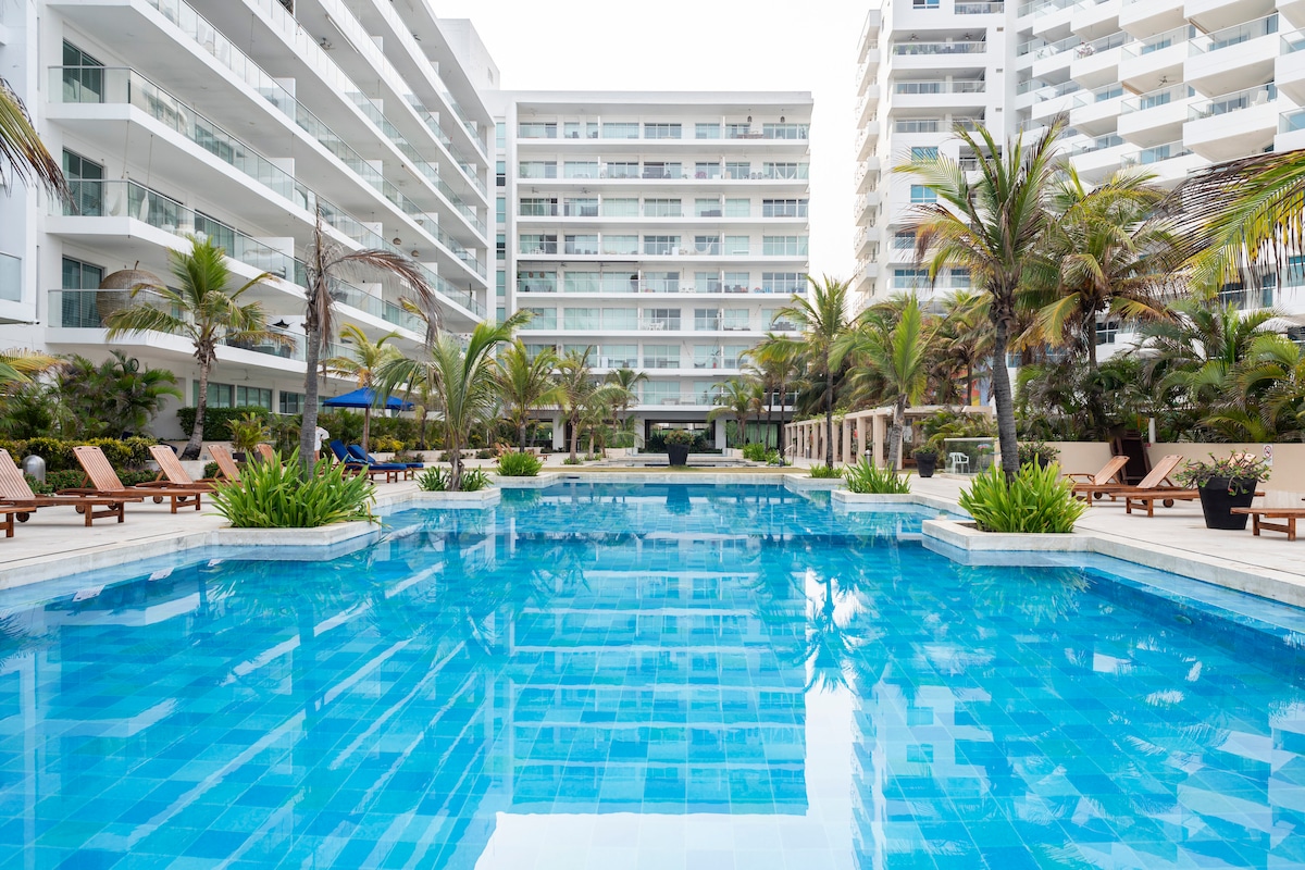 A large swimming pool reflects clear blue waters, surrounded by tropical palm trees and lounge chairs. The modern architecture of adjacent buildings adds a clean, contemporary touch to the serene outdoor space.