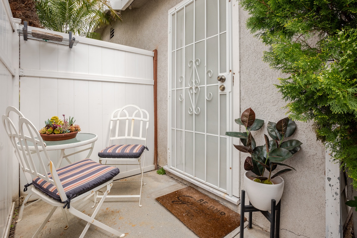 A small outdoor space is featured, containing two white chairs with striped cushions and a small round table. A welcome mat is positioned at the entrance door, surrounded by greenery, including a potted plant on a stand and a planter with a small succulent arrangement.