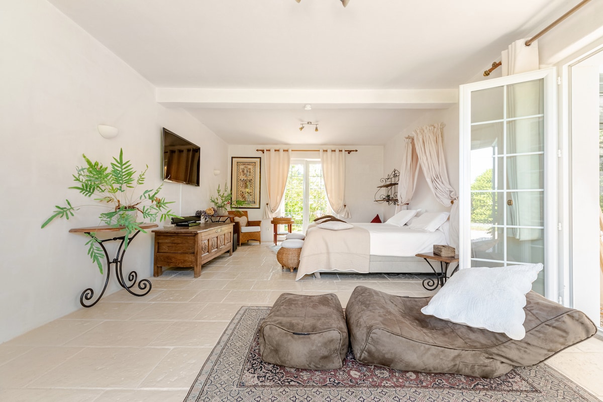 A spacious living area is presented, featuring a large bed and a soft chair, with natural light flooding through glass doors. A traditional rug adorns the floor, and a small table holds decorative elements, enhancing the serene ambiance of the space.