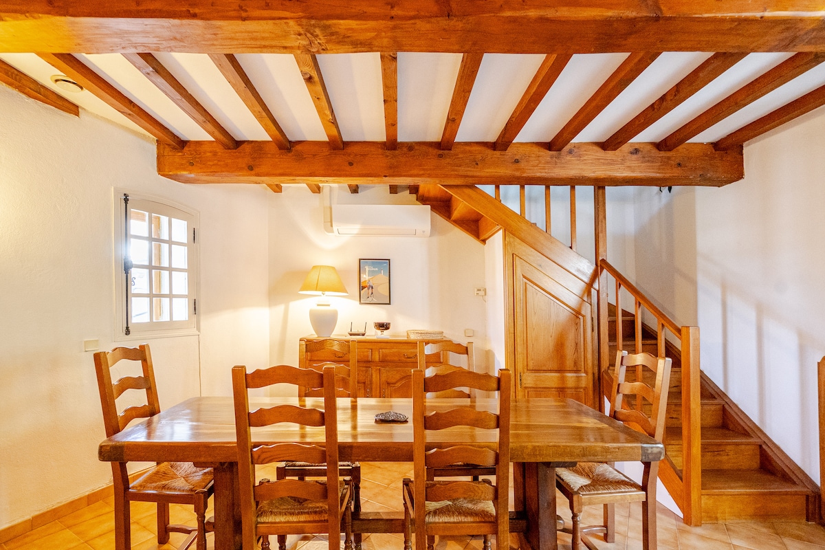 A spacious dining area features a wooden table surrounded by six wooden chairs. Exposed beam ceilings add character, and a staircase is visible in the background leading to the upper level. Soft light from a nearby lamp enhances the cozy ambiance of the room.