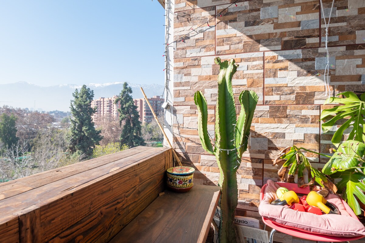 A wooden balcony railing is seen alongside a potted cactus and a leafy plant. A small table displays colorful decorative items. The backdrop showcases a clear blue sky and distant mountains, offering a peaceful view over the surrounding greenery.