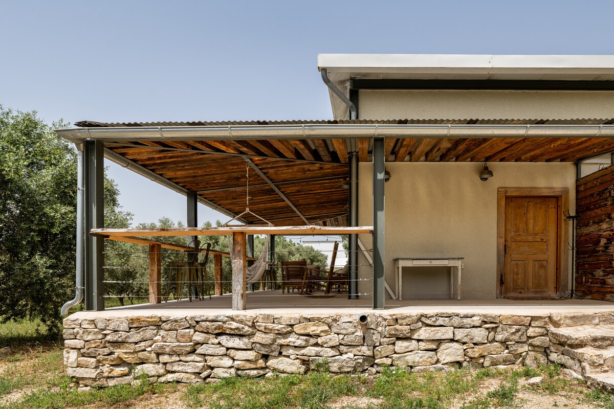 The outdoor area features a spacious pergola with wooden beams, offering shade and a relaxed atmosphere. A sturdy stone wall supports the structure, while a wooden door provides access to the interior. Casual seating and dining options are accommodated beneath the pergola.