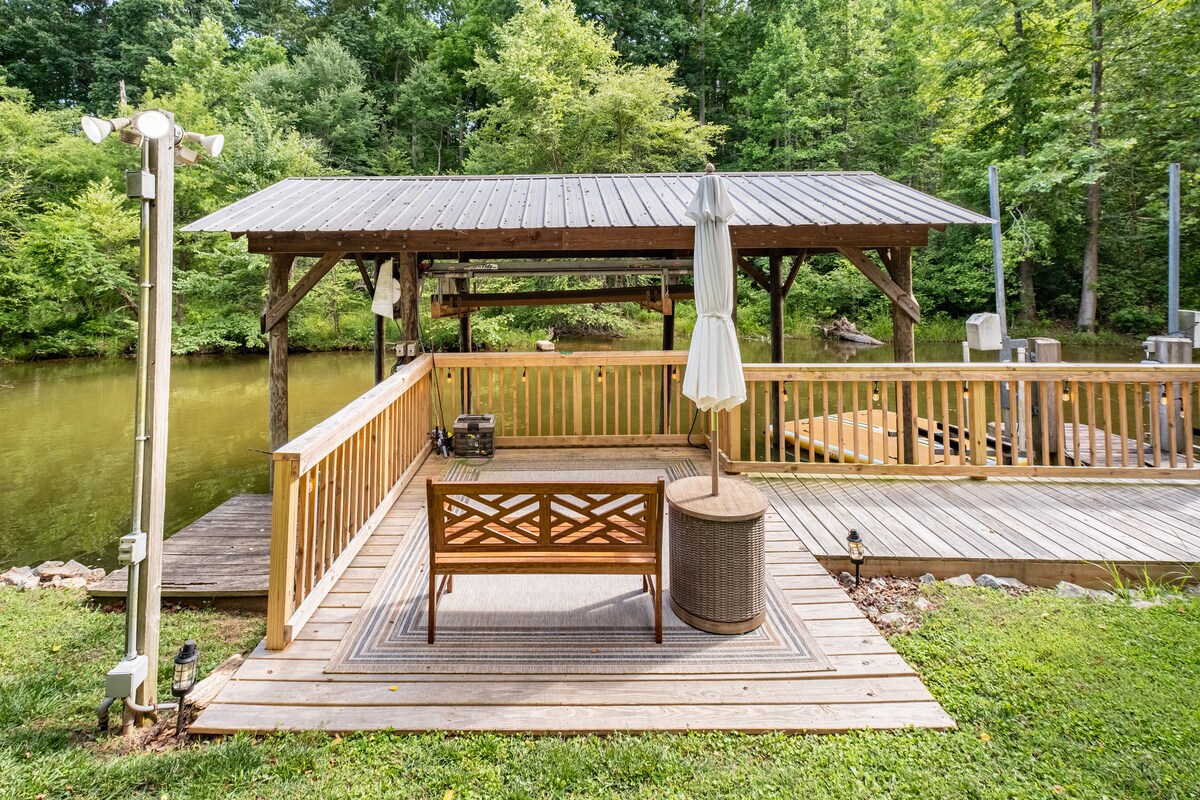 An outdoor deck space features a wooden railing and an umbrella, offering views of the calm lake surrounded by trees. Comfortable seating is provided for relaxation, with a rug enhancing the area. The serene setting invites guests to enjoy the natural surroundings.