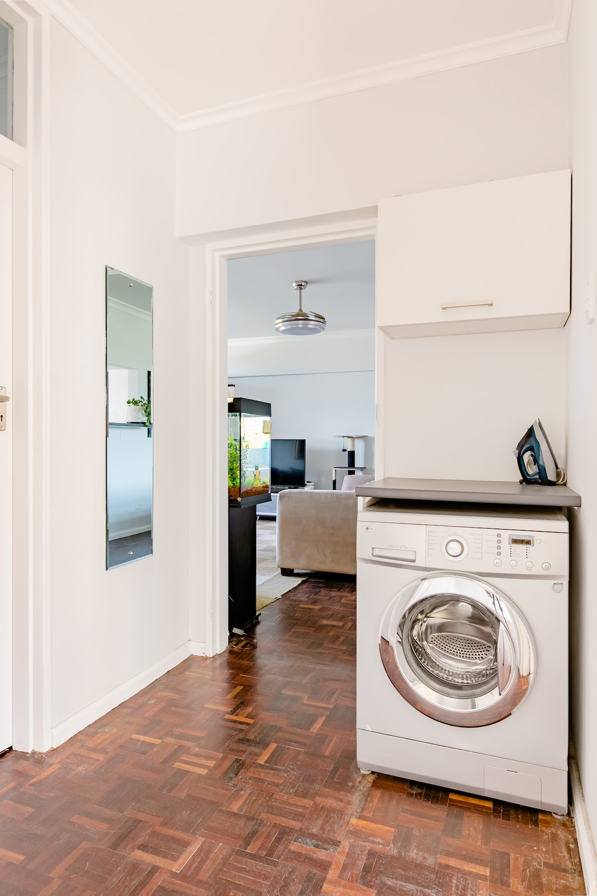 A compact laundry area features a front-loading washing machine and a sleek countertop. A large mirror reflects light, enhancing the space's openness. The room connects to a living area with visible seating options, providing a seamless flow throughout the apartment.