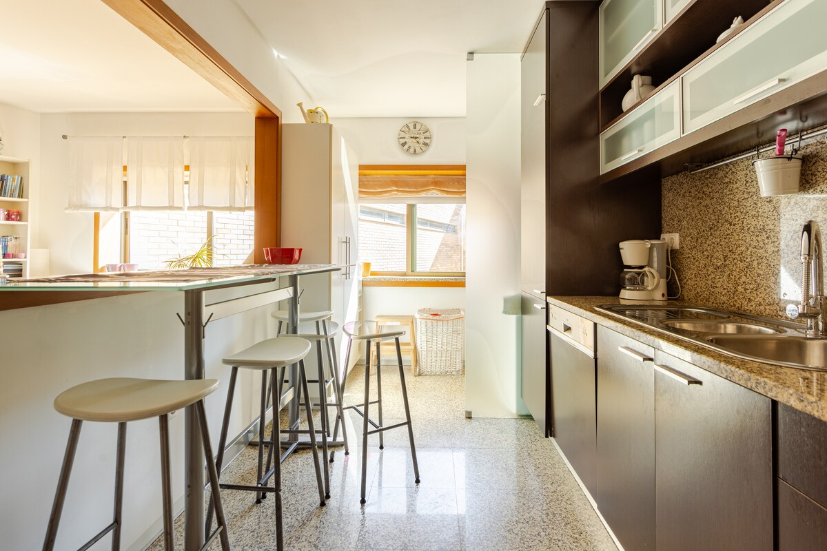 A modern kitchen is equipped with essential appliances and offers ample counter space. A breakfast bar with high stools provides a casual dining option. Natural light fills the room through a window, highlighting the sleek cabinetry and granite surfaces.