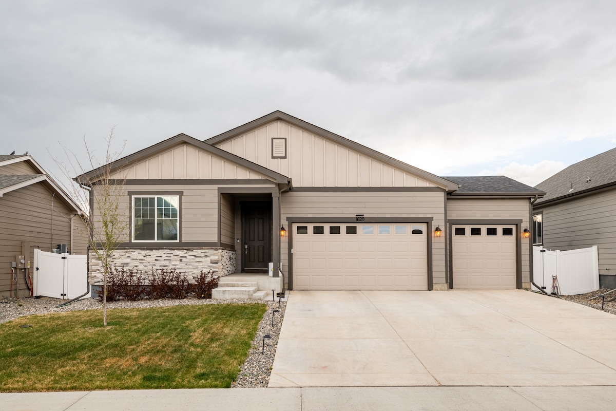 A modern two-story home is depicted with a light-colored exterior and a welcoming entrance. Two garage doors are visible, along with a neatly landscaped yard featuring grass and decorative stone elements. The surrounding area shows a suburban neighborhood setting.