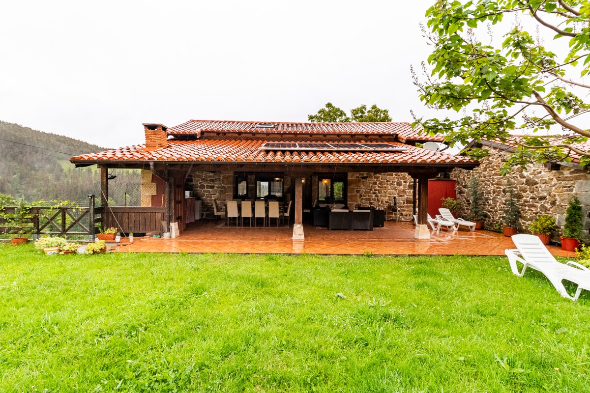 The exterior of a rustic rural home features a spacious wooden deck with several seating areas, overlooking a lush green lawn. Soft patio furniture is arranged for relaxation, while a backdrop of rolling hills is visible, suggesting a serene natural environment. A brick chimney adds charm to the structure.