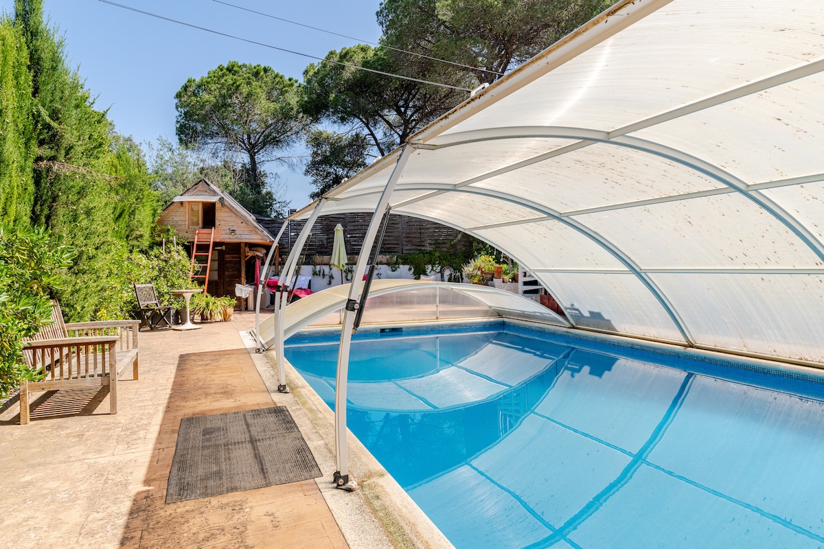 A covered swimming pool is displayed, reflecting clear blue water under a sunlit sky. Surrounding greenery contributes to a natural ambiance, while a wooden deck area offers seating options. A rustic chalet can be seen in the background, enhancing the outdoor space for relaxation.