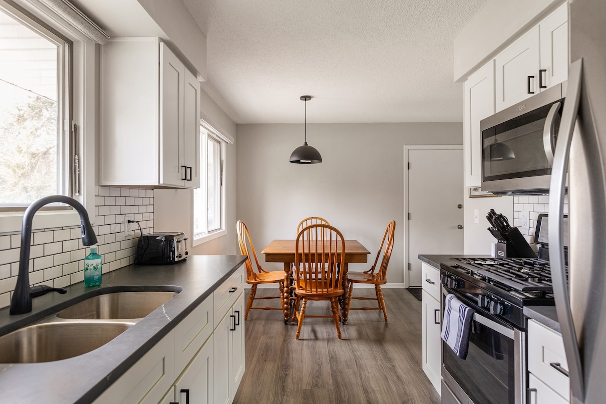A bright kitchen is showcased with modern white cabinetry and a gray backsplash. A dining area with a wooden table and four ladder-back chairs is visible, illuminated by natural light from the window. Stainless steel appliances are neatly arranged for convenience.