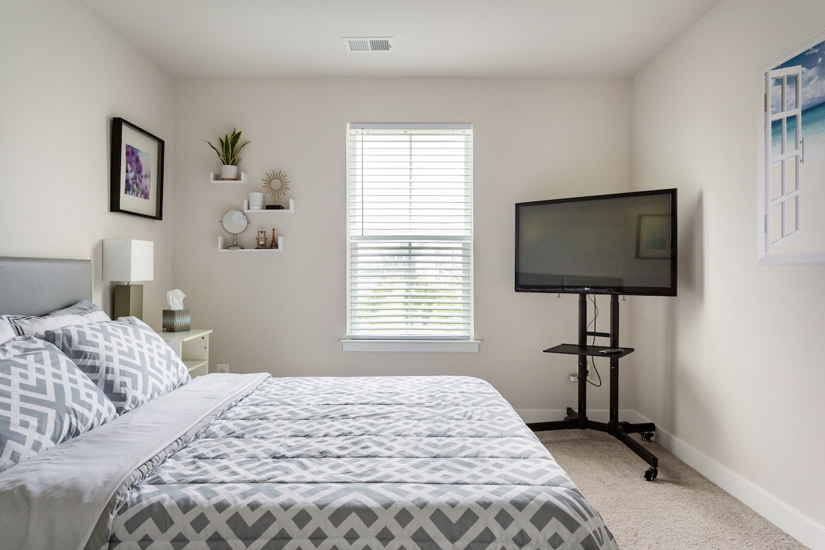 The room features a spacious bed with a geometric patterned bedding. A large window allows natural light to fill the space, framed by white blinds. On the wall, a flat-screen television is mounted beside a minimalist shelf displaying decorative items, while a small nightstand adds functionality.