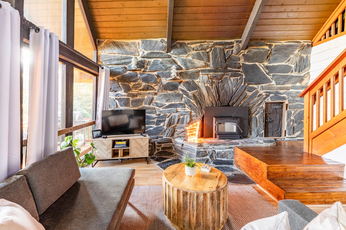 A welcoming living space features a striking stone fireplace surrounded by natural wood finishes. A cozy gray sofa and rustic wooden coffee table create an inviting area for relaxation, while expansive windows allow for ample natural light to fill the room.