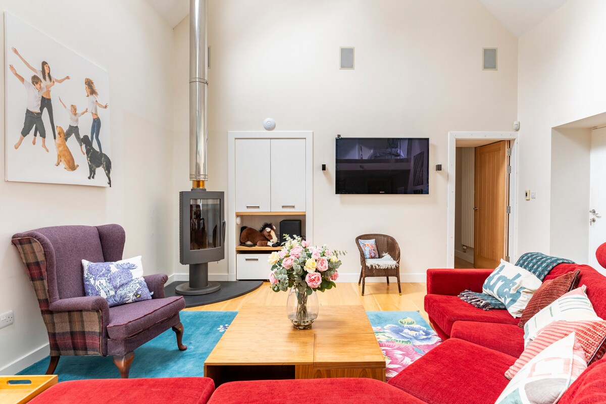 A spacious living area is characterized by a vibrant red sectional sofa and a cozy armchair. A wooden coffee table is set at the center, adorned with a floral arrangement. A contemporary wood-burning stove is positioned against the wall, paired with a wall-mounted television.