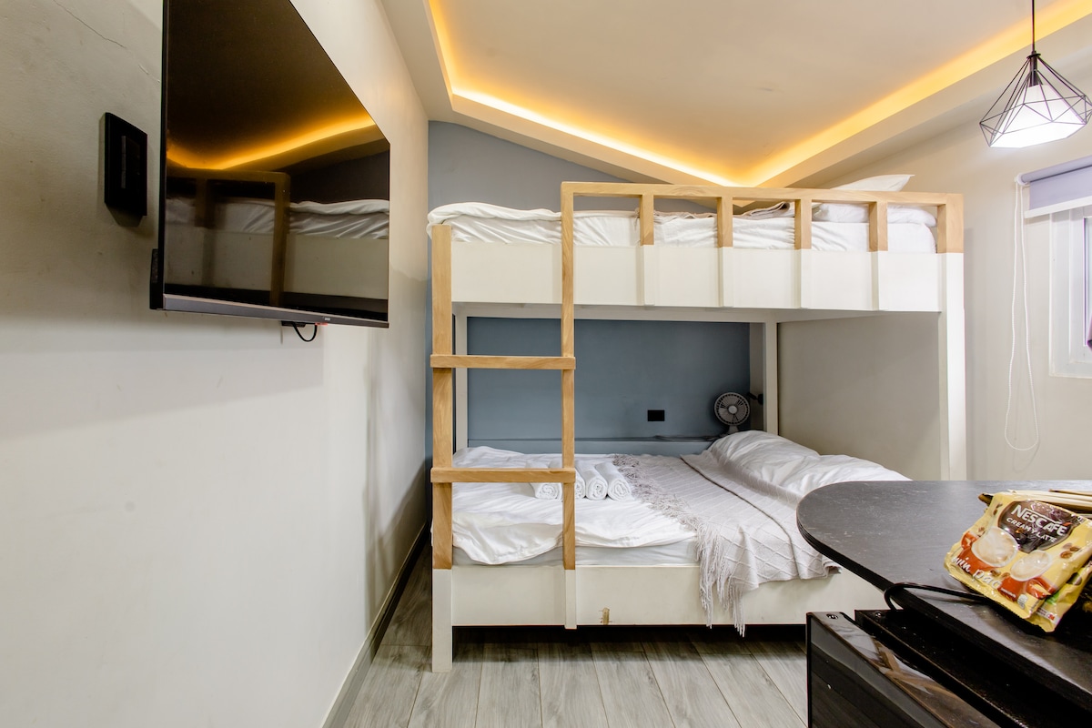 A modern sleeping area features a bunk bed with wooden framing, providing space for multiple guests. A television is mounted on the wall opposite the bed, while soft lighting is integrated into the ceiling. Bedding appears neat and inviting, enhancing the cozy environment.