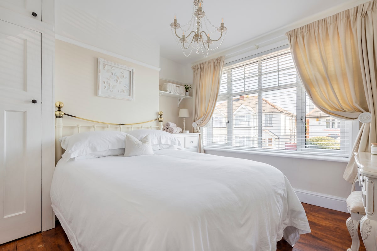 The inviting bedroom is furnished with a spacious bed dressed in crisp white linen. Natural light fills the room through large windows with sheer curtains. A chandelier hangs from the ceiling, complementing the warm tones of the decor and wooden flooring.