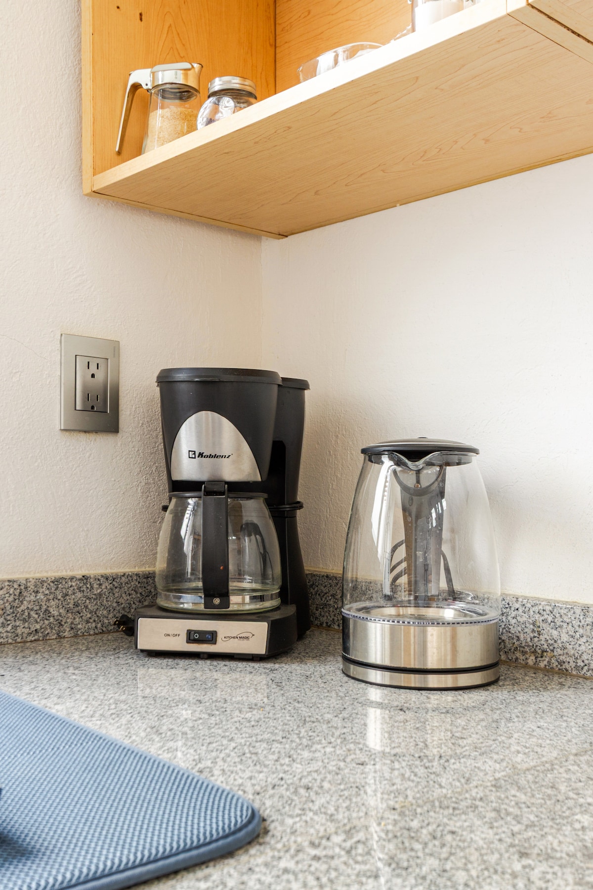 A corner of the kitchen features a coffee maker and a glass kettle on a granite countertop. Above, shelves hold jars and other kitchen items, providing accessible storage for essentials.