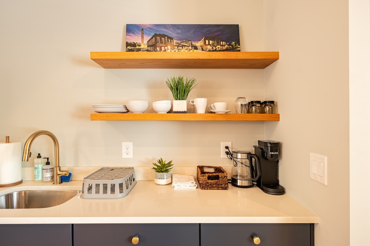 A well-organized kitchenette features sleek countertop surfaces and a sink. Wooden shelves display an array of dishware, including bowls and cups, alongside a coffee maker and a basket for additional storage. Natural greenery adds a touch of freshness to the space.