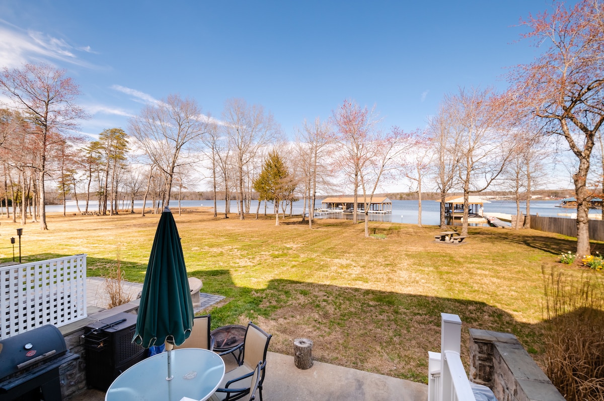 A serene lakeside view captures the expansive grassy area leading to the water. A patio table with chairs and a shaded umbrella is visible, alongside a charcoal grill. Dock access is seen in the distance, framed by leafless trees against a clear blue sky.