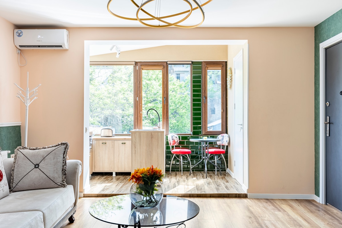 A modern living area is featured, showcasing a neutral-toned sofa and a circular glass coffee table. A decorative arrangement of flowers sits on the table. Beyond, a kitchen is visible with wooden cabinetry and a dining space accented by bright red chairs, framed by expansive windows overlooking greenery.