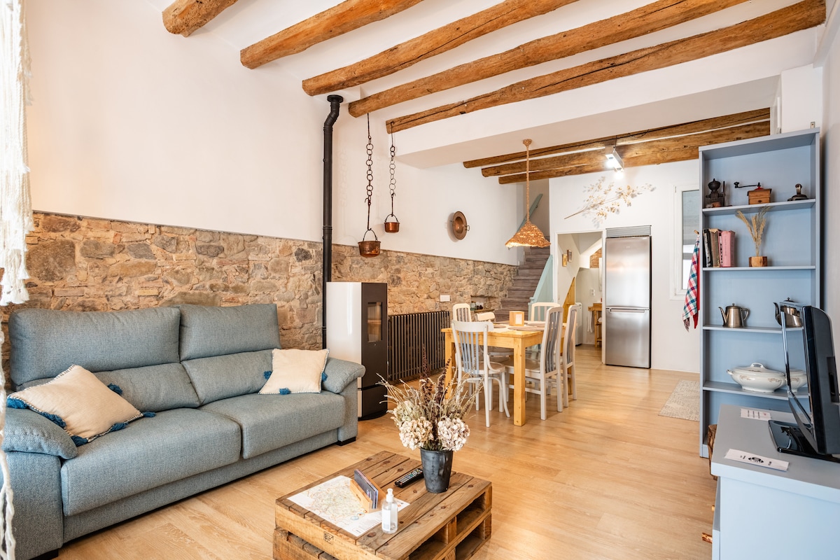 A spacious living area is highlighted by exposed wooden beams and a mix of stone and white walls. A large blue sofa faces a rustic coffee table, while a dining area with white chairs and a wooden table is set nearby. A sleek kitchen is partially visible in the background.