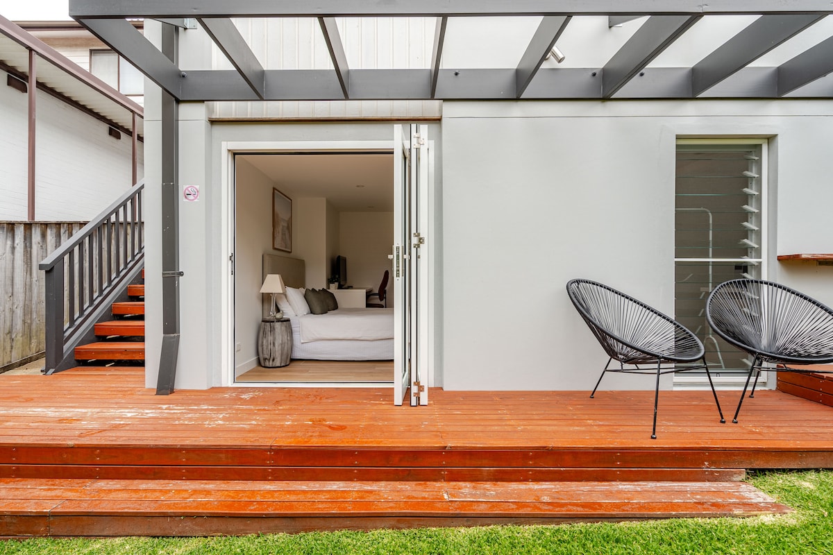The exterior features a private deck made of wood, leading to the studio apartment through large glass doors. Two black chairs are positioned on the deck, complementing the serene garden space visible beyond.