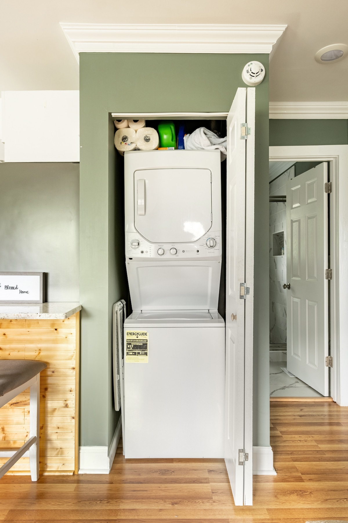 A compact laundry area is featured, showcasing a stacked washer and dryer within a closet space. Above, neatly arranged supplies, including paper towels, are visible. The space is enclosed by white doors, and a light-colored wooden surface is seen in the foreground.