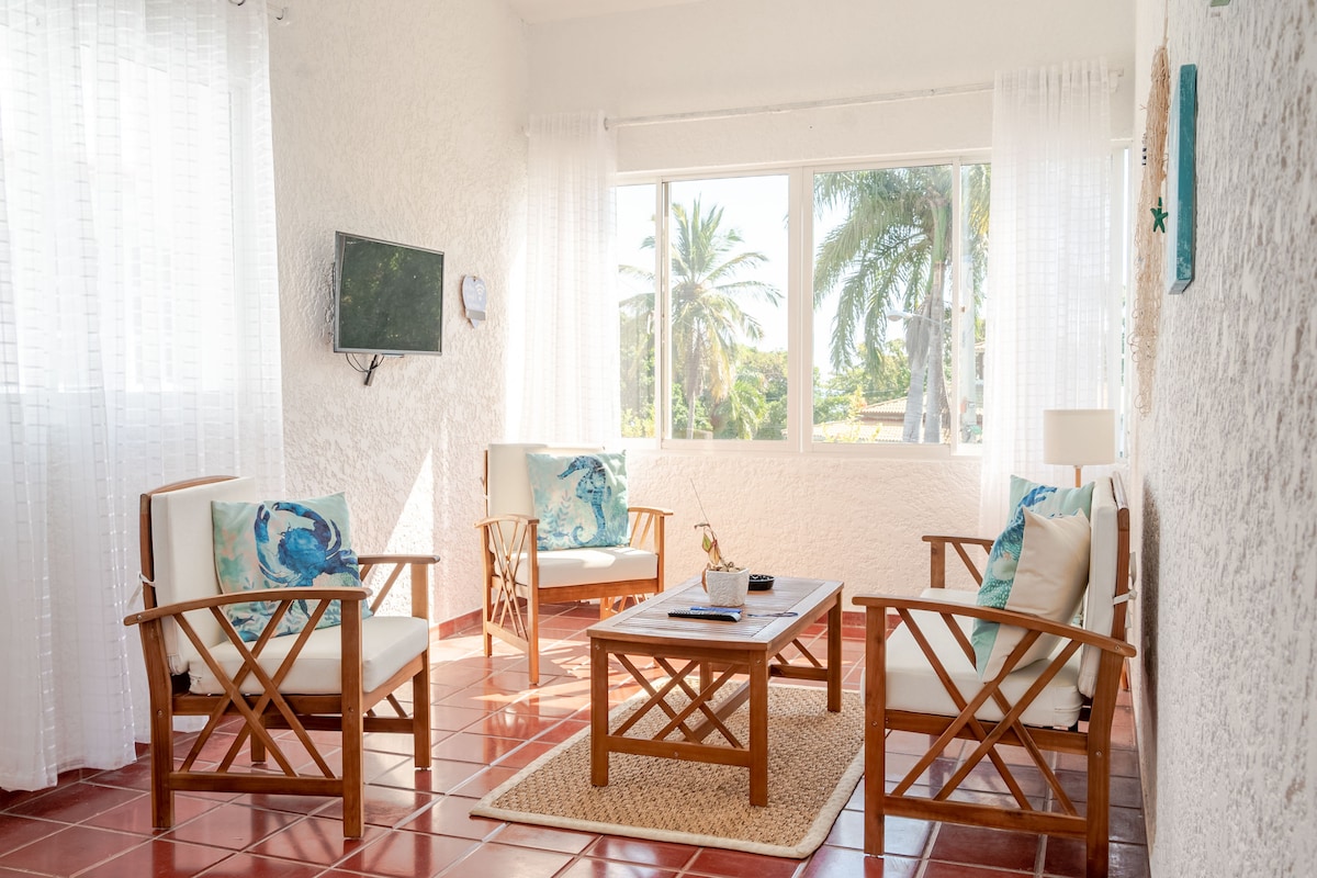 A bright living area features light-colored furniture with decorative cushions. A wooden coffee table is centered on a natural fiber rug. Large windows provide views of palm trees and allow natural light to fill the space.