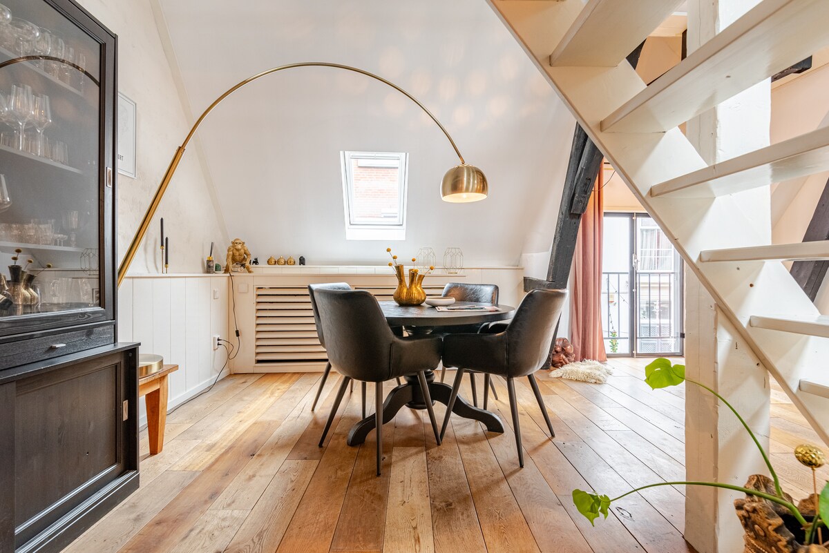A dining area is featured with a round table surrounded by four dark upholstered chairs. A modern arch lamp hangs above the table, and a side cabinet displays glassware. Natural light enters through a small window, highlighting the wooden floor.