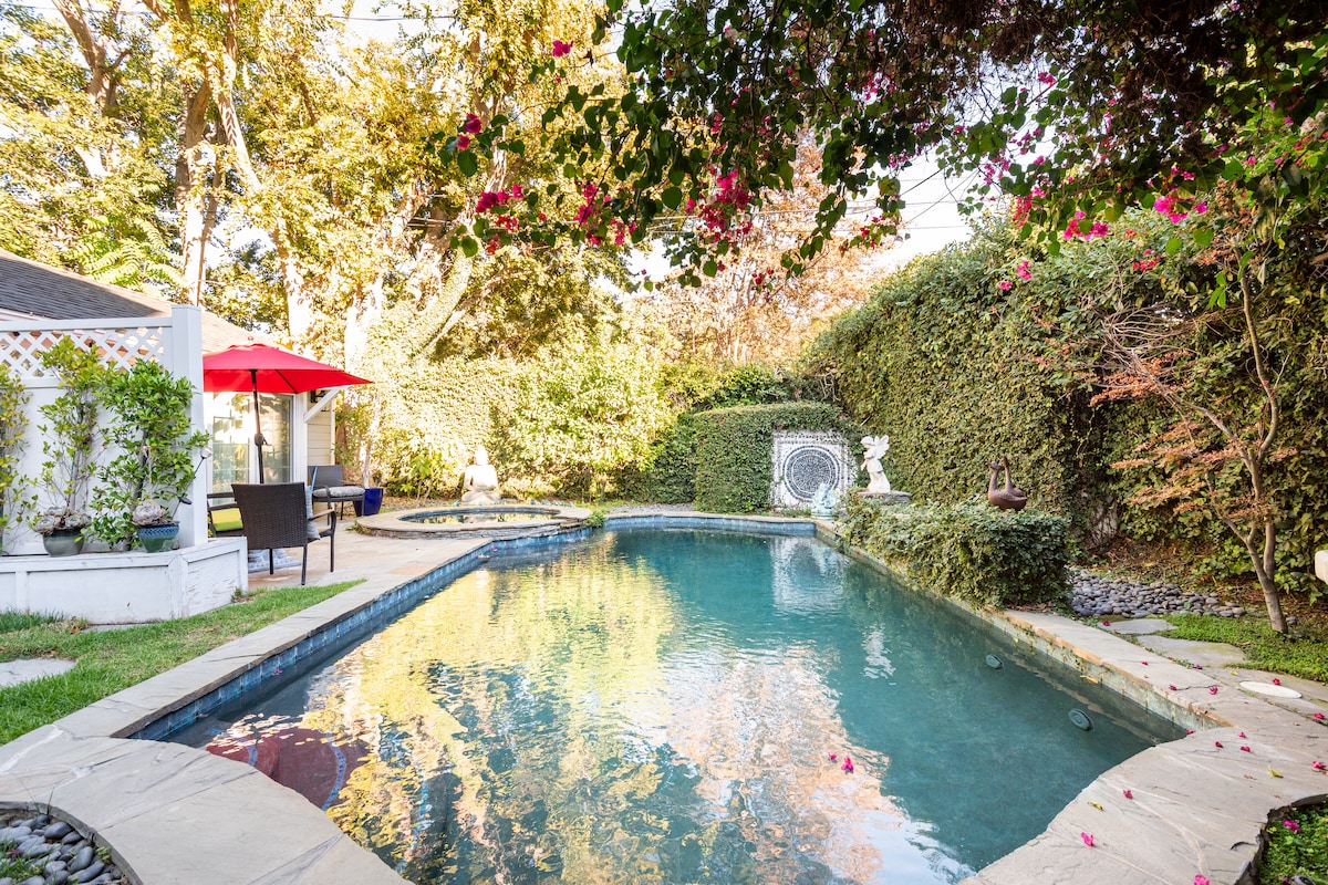 A serene backyard features a private pool surrounded by lush greenery and colorful flowering plants. A red umbrella shades sitting areas with dark wicker chairs near the water, creating a peaceful environment for relaxation. The space is enhanced by stone pathways and gentle landscaping.