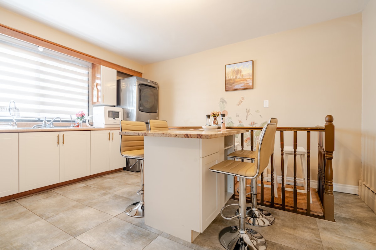 A light-filled kitchen area is highlighted by a central island with high stools. Light wood cabinetry is complemented by a beige wall, while a framed artwork adds a touch of decor. Large windows allow natural light to illuminate the space, enhancing its inviting atmosphere.