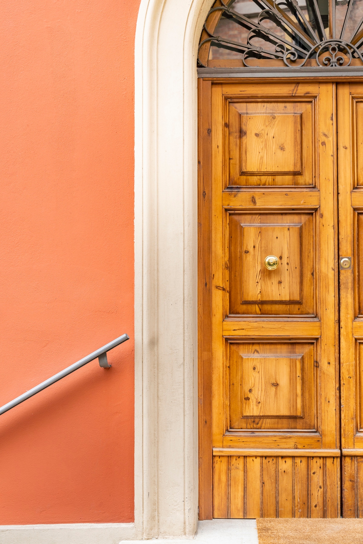 A wooden door with intricate paneling is framed by a soft, coral-colored wall. A decorative metal element crowns the door, while a handrail is positioned alongside the entrance, enhancing accessibility.