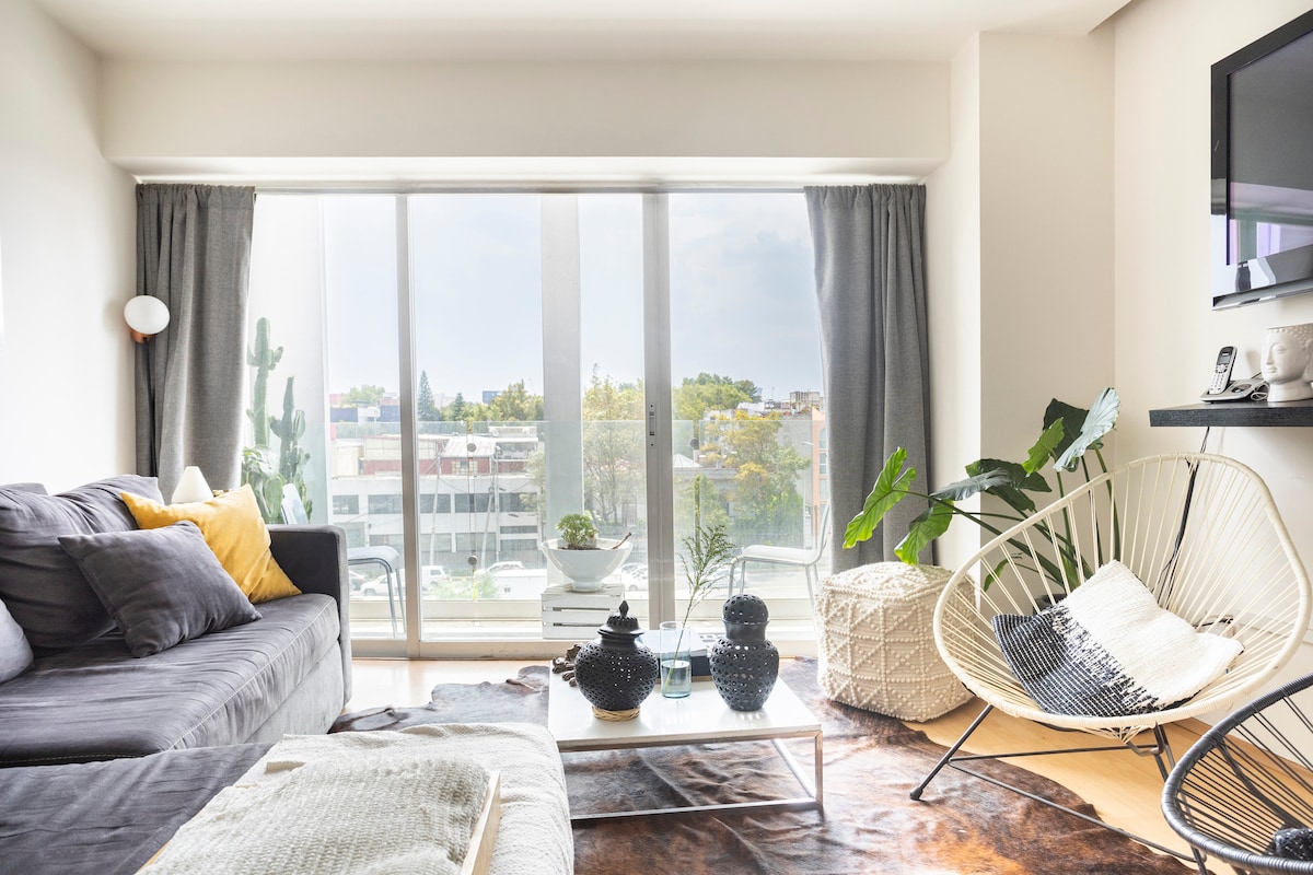 A bright living area features large glass windows allowing natural light to fill the space. A dark sofa is complemented by textured cushions and a cozy throw. An accent chair sits nearby, surrounded by decorative plants and various decorative objects on a glass table.