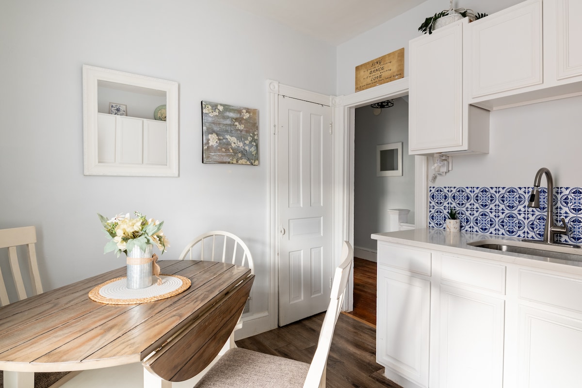 A light-filled eat-in kitchen is presented with a wooden dining table and white chairs. Blue patterned backsplash tiles add a pop of color above the kitchen sink. Decorative wall art enhances the space, complemented by a small vase of flowers on the table.