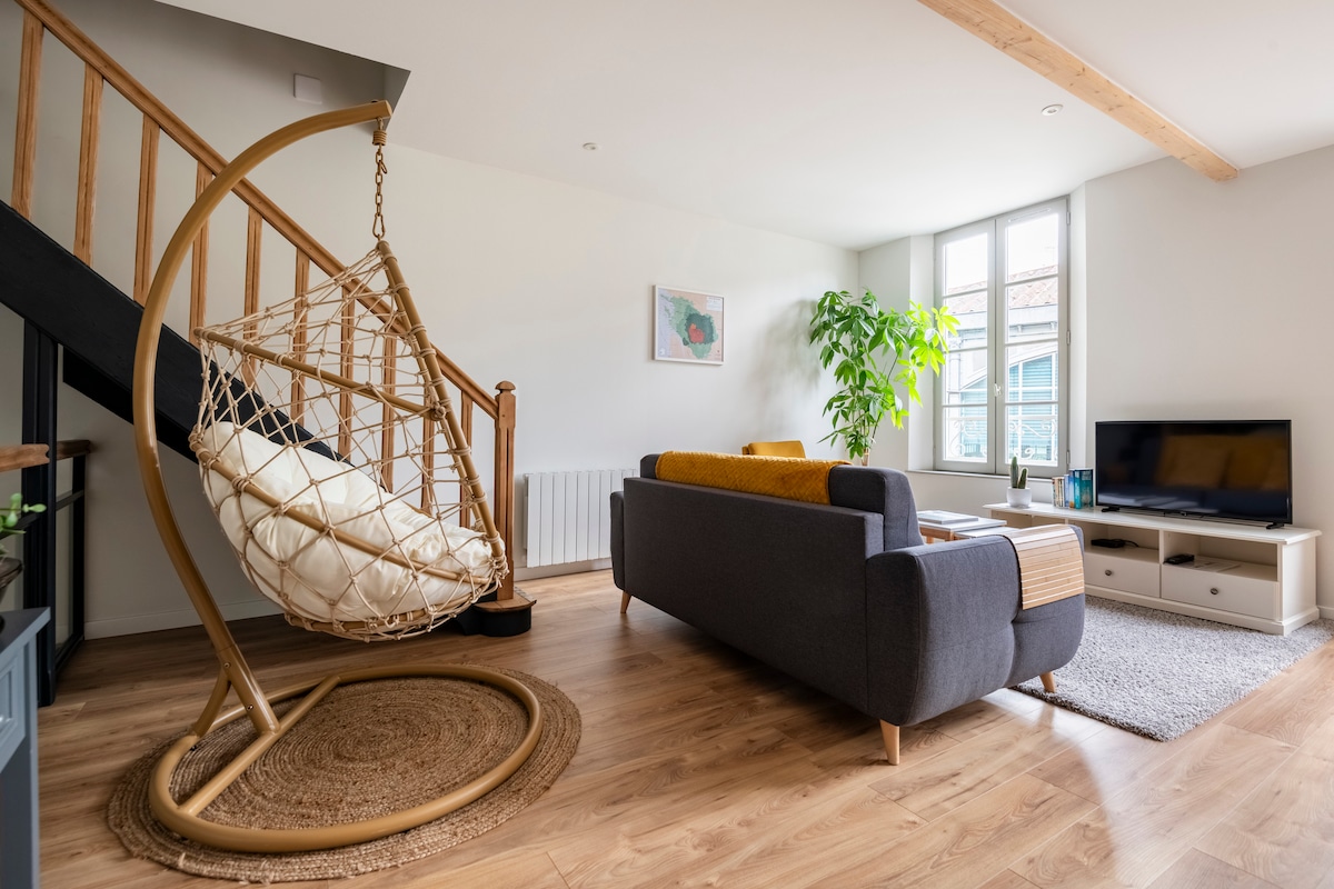 The living area features a cozy seating arrangement with a dark gray sofa and a bright yellow throw blanket. A hanging chair is positioned next to a set of stairs, while a potted plant adds a touch of greenery. Large windows provide natural light.